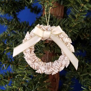 May include: A white seashell wreath ornament with a white satin ribbon with gold trim. The ornament is hanging on a green Christmas tree.