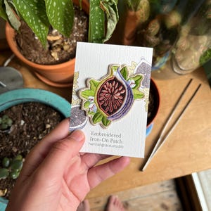 May include: Embroidered iron-on patch with a fig design in purple, green, and red. The patch is on a white card with the text "Embroidered Iron-On Patch hannahgrace.studio".