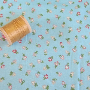Könnte beinhalten: Eine Spule beigefarbenen Fadens liegt auf einem hellblauen Stoff, der mit kleinen Blumenmustern in Rosa, Weiß und Grün bedeckt ist. Der Stoff hat eine strukturierte Oberfläche, die auf ein gewebtes Material hindeutet. Der Faden ist fest auf eine Holzspule gewickelt.