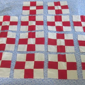 May include: A collection of fourteen square fabric quilt blocks arranged in a grid. Each block features a nine-patch pattern consisting of alternating red and off-white squares, creating a consistent checkerboard design across all individual pieces.