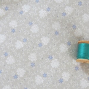 Puede incluir: Una bobina de hilo turquesa en un carrete de madera. El carrete está sobre una tela gris claro con un patrón floral blanco.