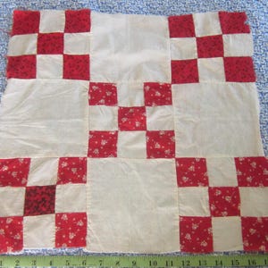 May include: A square fabric quilt block featuring a nine-patch pattern with alternating solid off-white squares and red floral print squares. A green measuring tape is positioned along the bottom edge, showing a width of approximately 19 inches.