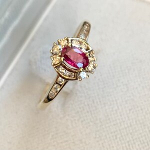 May include: A gold ring with an oval ruby centre stone, encircled by a halo of small diamonds. The ring's band is also set with diamonds. The jewellery is displayed on a white surface, accentuating its brilliance and design.