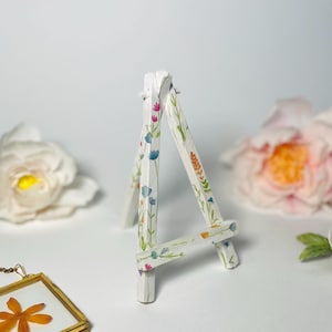 Puede incluir: Un pequeño caballete de madera blanco decorado con patrones florales coloridos. El caballete en miniatura está diseñado para sostener pequeñas obras de arte o artículos de exhibición. El diseño floral incluye flores azules, rosas y naranjas con hojas verdes.