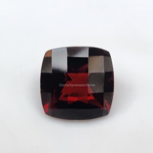 May include: A deep red gemstone with a cushion cut and a high polish. The gemstone is set in a clear, faceted setting. The gemstone is approximately 10mm x 8mm in size. GemsXpressionStore is printed on the gemstone.