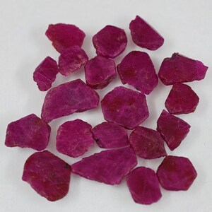 May include: A collection of small, raw, unpolished ruby gemstones with a deep magenta-red color. Each stone features irregular, jagged edges and a matte, textured surface, showcasing natural crystalline formations in various shapes and sizes.