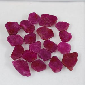 May include: A collection of small, raw, unpolished ruby gemstones with irregular shapes and varying shades of deep magenta and vibrant pink. Each stone features a textured, matte surface with natural facets and edges scattered across the frame.