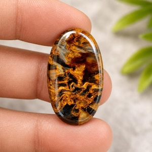 May include: Oval-shaped gemstone with a swirling pattern of gold, brown, and black. The polished surface reflects light, highlighting the intricate natural design. This gemstone is a unique piece for jewelry making.