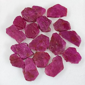 May include: A collection of fifteen raw, unpolished ruby gemstones with a vibrant magenta-pink hue. Each stone features an irregular, jagged shape with a textured, crystalline surface, showcasing natural facets and deep, saturated color variations.