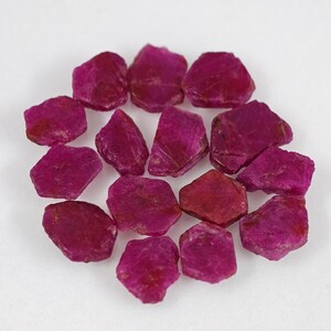 May include: A collection of fifteen loose, raw ruby gemstones. Each stone features an irregular, multifaceted shape with a deep magenta-red color and a natural, unpolished texture. The rubies are arranged in a circular cluster on a plain surface.