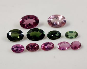 May include: Assortment of oval-shaped gemstones in shades of pink, green, and black. The gemstones are arranged in a row on a white surface.