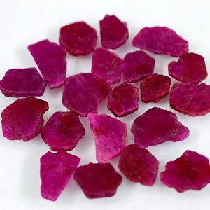 May include: A collection of small, raw, unpolished ruby gemstones in a vibrant deep pinkish-red hue. Each stone features irregular, jagged edges and a natural, textured surface with varying levels of opacity and crystalline structure throughout.