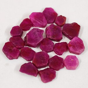 May include: A collection of small, raw, unpolished ruby gemstones in various shades of deep pink and magenta. Each stone features an irregular, multifaceted shape with a matte, textured surface, scattered together in a loose cluster.