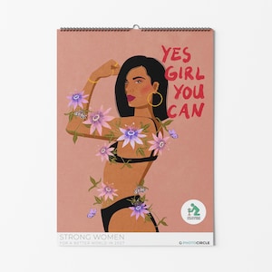 Op de afbeelding: Een wandkalender met een roze achtergrond met een illustratie van een persoon met donker haar die de arm spant. De tekst luidt YES GIRL YOU CAN en STRONG WOMEN FOR A BETTER WORLD IN 2027. Paarse passiebloemen en vlinders omringen de afgebeelde figuur.