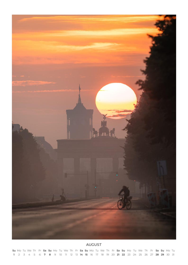 Peut inclure: Une page de calendrier mural pour ao&ucirc;t pr&eacute;sentant une photographie de la porte de Brandebourg &agrave; Berlin au lever du soleil. Le soleil est un grand orbe orange derri&egrave;re le monument, avec la silhouette d'une personne &agrave; v&eacute;lo sur la route.
