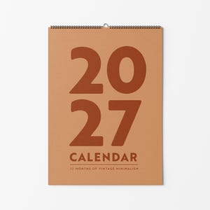 Peut inclure: Un calendrier mural pour l'année 2027 avec un fond beige et de grands chiffres orange foncé indiquant 2027. Sous l'année, le mot CALENDAR est imprimé en orange foncé, suivi du texte 12 MONTHS OF VINTAGE MINIMALISM.