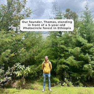 Peut inclure: Une personne portant un sweat &agrave; capuche jaune et un jean bleu se tient devant une for&ecirc;t verte dense. Un panneau rectangulaire blanc avec du texte noir indique : Our founder, Thomas, standing in front of a 5-year-old Photocircle forest in Ethiopia.