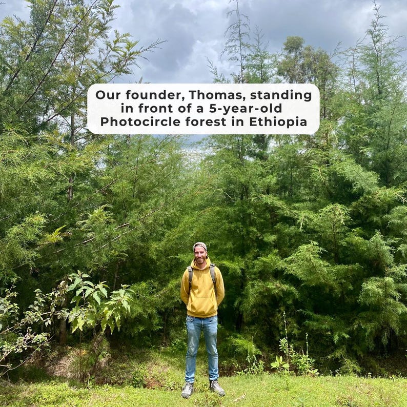 Peut inclure: Une personne portant un sweat &agrave; capuche jaune et un jean bleu se tient devant une for&ecirc;t verte dense. Un panneau rectangulaire blanc avec du texte noir indique : Our founder, Thomas, standing in front of a 5-year-old Photocircle forest in Ethiopia.