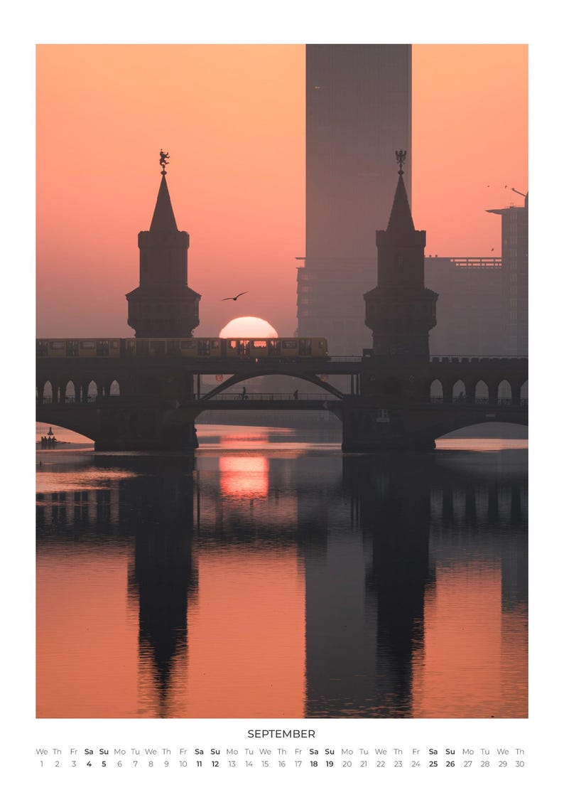 Peut inclure: Une page de calendrier pour septembre pr&eacute;sentant une photographie du pont Oberbaum &agrave; Berlin au lever du soleil. La silhouette du pont et d'un train se refl&egrave;tent dans l'eau sous un ciel orange vif. La grille du calendrier est imprim&eacute;e en bas.