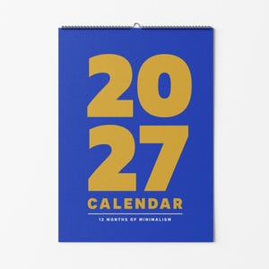 Könnte beinhalten: Ein Wandkalender mit blauem Hintergrund und großen goldenen Ziffern 2027 in der Mitte. Unter dem Jahr steht das Wort CALENDAR in Gold, gefolgt von dem Text 12 MONTHS OF MINIMALISM in Weiß, alles zusammengehalten durch eine schwarze Drahtspirale.