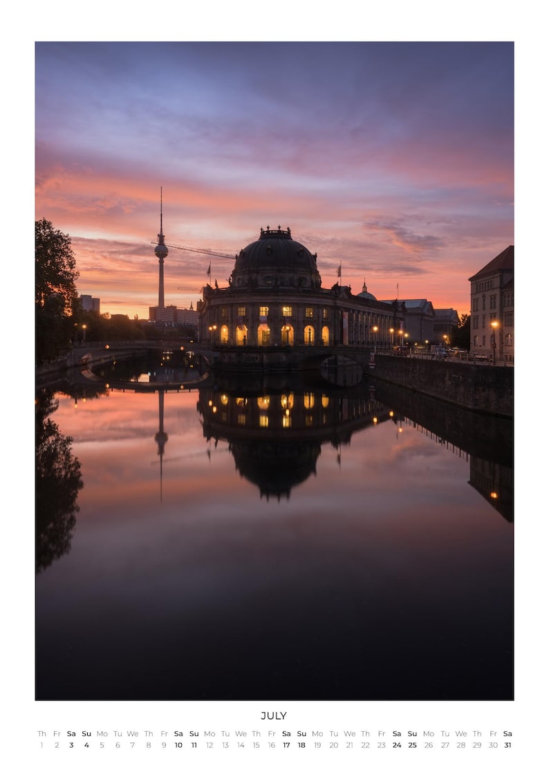 Peut inclure: Une page de calendrier pour juillet pr&eacute;sentant une photographie du mus&eacute;e Bode &agrave; Berlin au coucher du soleil, avec la tour de t&eacute;l&eacute;vision visible au loin. Le b&acirc;timent est illumin&eacute; et se refl&egrave;te dans l'eau calme sous un ciel rose et orange.