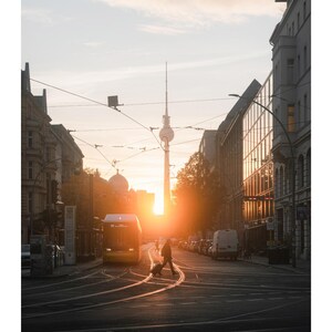 Peut inclure: Une page de calendrier mural pour mai pr&eacute;sentant une photographie d'une rue de Berlin au coucher du soleil. La tour de t&eacute;l&eacute;vision embl&eacute;matique se dresse au loin, encadr&eacute;e par des b&acirc;timents, des rails de tramway, un tram et un pi&eacute;ton avec bagages.