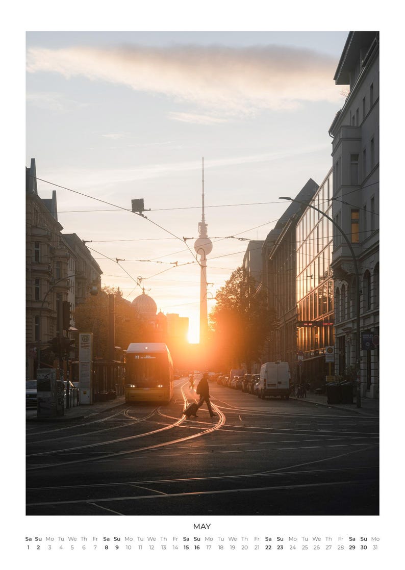 Peut inclure: Une page de calendrier mural pour mai pr&eacute;sentant une photographie d'une rue de Berlin au coucher du soleil. La tour de t&eacute;l&eacute;vision embl&eacute;matique se dresse au loin, encadr&eacute;e par des b&acirc;timents, des rails de tramway, un tram et un pi&eacute;ton avec bagages.