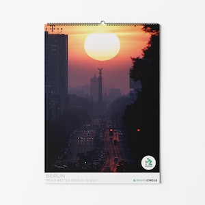 Peut inclure: Un calendrier mural pr&eacute;sentant une vue au coucher du soleil de la colonne de la Victoire de Berlin et de l'horizon urbain. L'image montre un grand soleil derri&egrave;re le monument, avec une route et du trafic. Texte : BERLIN FOR A BETTER WORLD IN 2027.