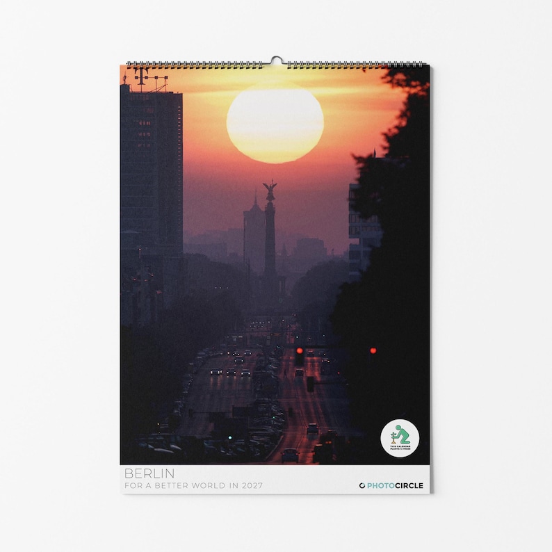 Peut inclure: Un calendrier mural pr&eacute;sentant une vue au coucher du soleil de la colonne de la Victoire de Berlin et de l'horizon urbain. L'image montre un grand soleil derri&egrave;re le monument, avec une route et du trafic. Texte : BERLIN FOR A BETTER WORLD IN 2027.