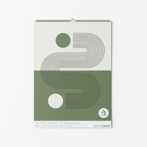 Puede incluir: Un calendario de pared con encuadernación en espiral, con un diseño minimalista en verde oliva y blanco roto. El diseño incluye líneas negras curvas y dos círculos. El texto dice: "IN THE SPIRIT OF BAUHAUS FOR A BETTER WORLD IN 2026."