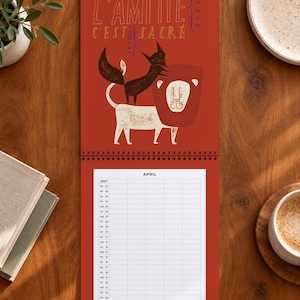 Peut inclure: Un calendrier mural pour avril 2027 sur fond rouge illustré d'un renard noir debout sur un lion blanc. Le texte en haut indique L'AMITIÉ C'EST SACRÉ et VRAIMENT AMOUR. La grille du calendrier en dessous liste les jours du mois verticalement.