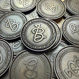 May include: A collection of circular antique bronze-toned metal coins featuring a raised monogram of the letters JB in the center. The text 2026 BOGEY BROS INVITATIONAL SAINT GEORGE, UTAH is embossed in a circular pattern around the edge of each coin.