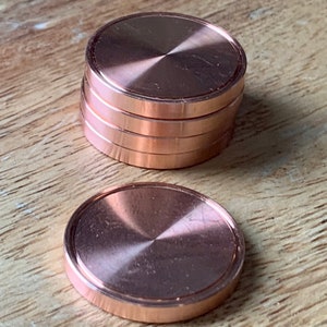 以下が含まれることがあります： 銅色の丸い金属製のコースターが5つ積み重ねられています。コースターは滑らかで光沢のある表面とわずかに盛り上がった縁を持っています。