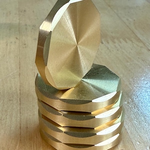 Op de afbeelding: Een stapel van zes goudkleurige, facetgeslepen metalen schijven, met één schijf iets uit het midden. De schijven hebben een geometrisch ontwerp en een gepolijste afwerking, die licht reflecteert. De achtergrond is een lichtgekleurd houten oppervlak.