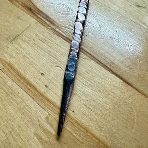 May include: A handcrafted copper tool with a textured, hammered design. The tool has a flat, rectangular top with circular indentations, tapering to a pointed end. The copper has a dark, aged patina, and it rests on a wooden surface.