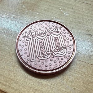 May include: A circular copper-toned commemorative coin featuring the embossed text JOHN MOORE 100 LESSONS. The surface is decorated with a repeating pattern of small four-pointed stars, and the coin has a textured, ridged edge around its perimeter.