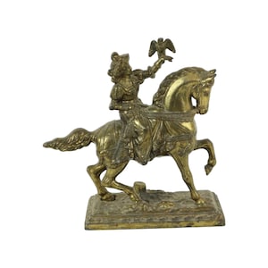 May include: A gold-toned metal figurine of a woman riding a horse. The woman is holding a bird in her right hand. The figurine is on a rectangular base.
