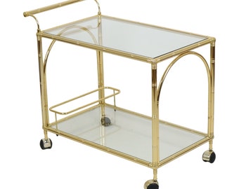 Könnte beinhalten: Ein goldfarbener Barwagen aus Metall mit zwei Glasböden und Rädern. Der Wagen hat einen goldfarbenen Metallgriff und einen goldfarbenen Metallrahmen.