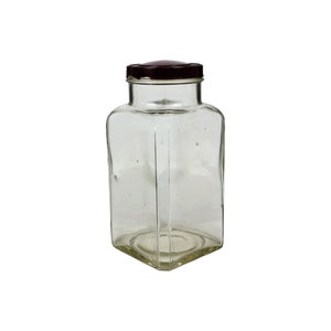 May include: A clear glass jar with a square base and a dark brown lid. The jar has a rectangular body with rounded corners. The lid is a solid, dark brown color. The jar is empty and appears to be vintage.
