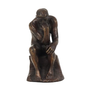 Op de afbeelding: Een bronskleurig beeldje van een zittende figuur in een contemplatieve houding, met de kin rustend op een hand. Het figuur heeft een getextureerde, metalen afwerking en is afgebeeld in een gebogen houding op een rotsachtige basis.