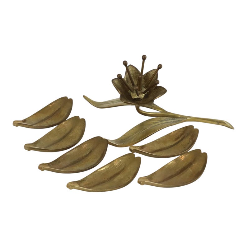 Op de afbeelding: Een messing set bestaande uit een centraal bloemsculptuur met een lange steel en zes afzonderlijke bladvormige schaaltjes. Elk stuk heeft een getextureerde, metallic gouden afwerking met zichtbare nerven op de bladeren en meeldraden.