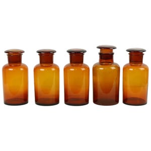 May include: Five amber glass bottles with glass stoppers. The bottles are all the same size and shape, and they are arranged in a row.