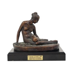 May include: A bronze sculpture of a nude woman in a seated position, with her head bowed. The sculpture is mounted on a black rectangular base. The text on the base reads "La Nymphe au scorpion - 1835 Reproduction en bronze D'après l'oeuvre de Bartolini."