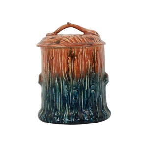 May include: A ceramic jar shaped like a tree stump with a lid. The jar is painted in shades of brown, blue, and green to resemble wood grain.
