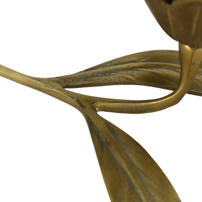 Op de afbeelding: Een koperen kandelaar in de vorm van een bloem met een steel en twee bladeren. Het metaal heeft een warme, goudbruine tint met een licht verweerde, getextureerde afwerking. Het ontwerp toont een gebogen steel die uitloopt in twee bladeren.