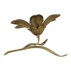 Op de afbeelding: Een koperen kandelaar in de vorm van een bloeiende bloem met brede bloemblaadjes en een centrale houder voor een kaars. De basis heeft een gebogen steel en twee lange, slanke bladeren die naar buiten uitsteken, wat een gebalanceerd metalen object vormt.