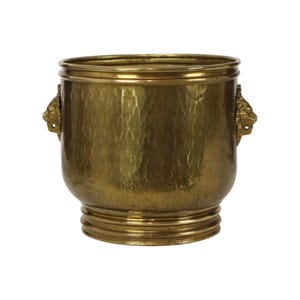 May include: A hammered brass bucket with a polished finish, featuring a wide circular rim, a tiered base, and two decorative side handles shaped like lion heads. The metal surface displays a textured, dimpled pattern across the exterior body.