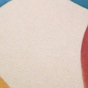 May include: A close-up of a cream-colored fabric with a subtle texture. The fabric is part of a larger design with a red, orange, and blue color scheme.