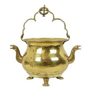 Pode incluir: Um caldeirão de latão vintage com uma alça decorativa e bicos em forma de pássaro. O caldeirão tem três pés e uma textura martelada, dando-lhe uma aparência antiga. A alça tem um design ornamentado.
