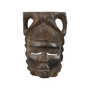 May include: A dark brown wooden mask with a carved human face. The mask has a detailed design with a raised forehead and a carved crown. The eyes are closed and the mouth is slightly open.